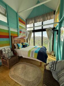 Queen Bed with white sheets and blue and green comforter, with colorful wooden walls and 2 wicker chairs with accented blankes and pillows