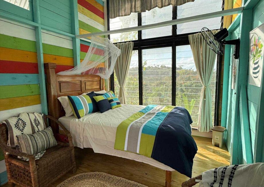 Queen Bed with white sheets and blue and green comforter, with colorful wooden walls and 2 wicker chairs with accented blankes and pillows