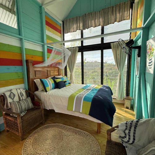 Queen Bed with white sheets and blue and green comforter, with colorful wooden walls and 2 wicker chairs with accented blankes and pillows
